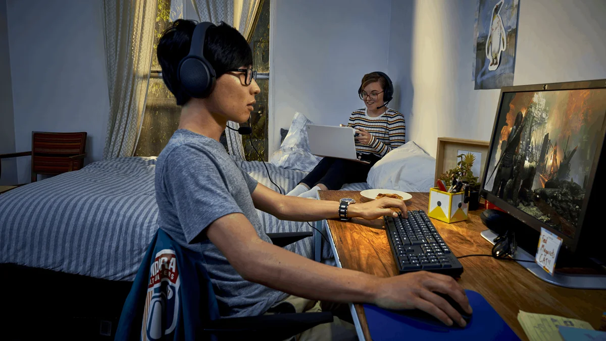 Two college students sit in their dorm playing Stadia games on their computers.