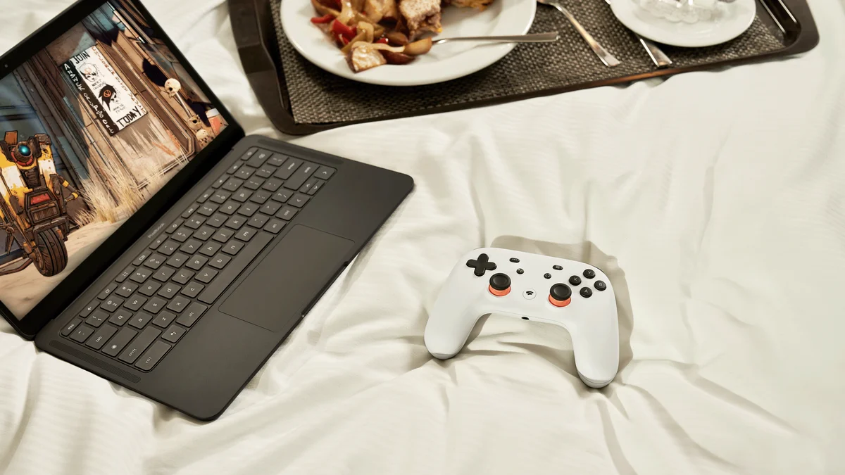 A white Stadia controller sits on a white bed with a black laptop showing a game.