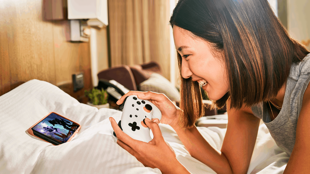A woman with brown hair holds a white Stadia controller on a white bed, looking at her phone as she plays a game on Stadia.
