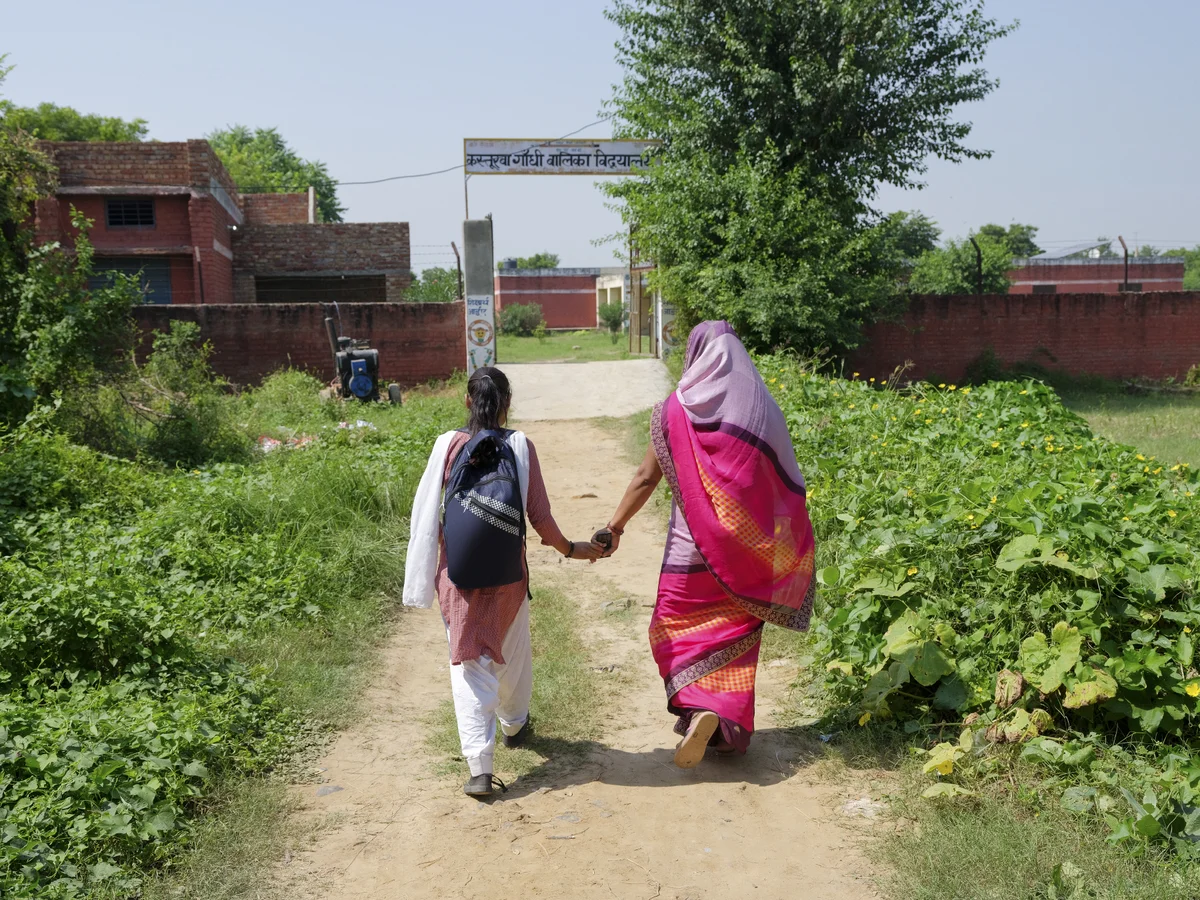 Two women, one carrying a backpack and the other dressed in a pink and purple sari, walk hand-in-hand down a pathway toward a building, with greenery on either side.