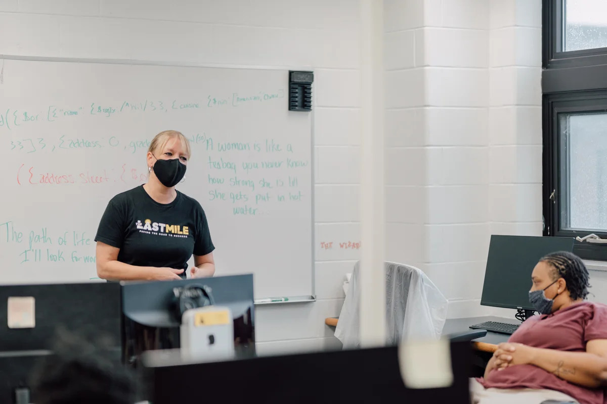 A woman wearing a mask, standing in front of a whiteboard, teaching a digital skills class at The Last Mile, a nonprofit partner who supports training formerly incarcerated individuals.