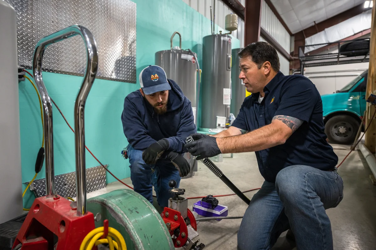 Two members of the team at A&A Plumbing, wearing dark blue shirts and jeans, discuss a piece of equipment in a workshop. A truck is visible in the background.