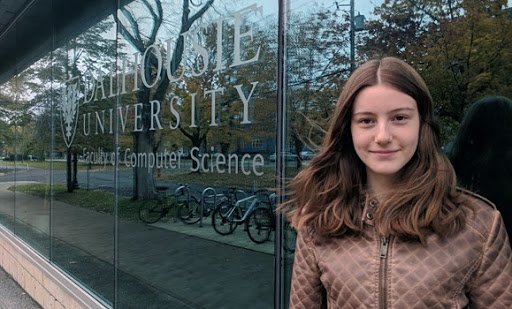 Image shows a young woman with brown hair wearing a brown coat, looking into the camera and smiling. She's standing against a window to a building that says "Dalhousie University, department of Computer Science."