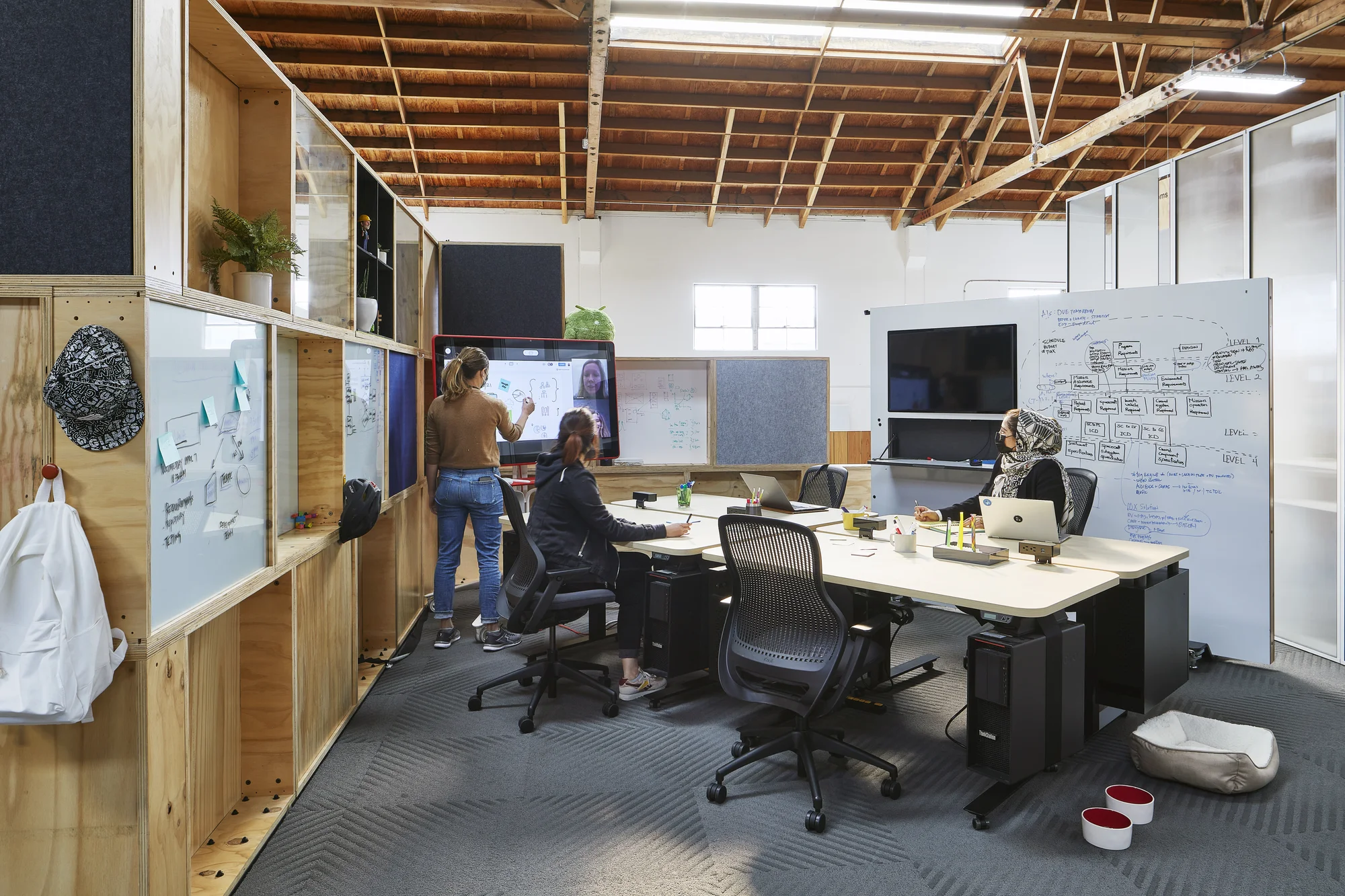 A "Team Pod" with a movable whiteboard wall, 4 desks on rollers and other reconfigurable furnishings, with 2 people sitting at desks and 1 person standing at a Jamboard, with another person on the videoconference