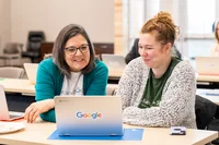 Two women are seated together at a table smiling as they review information on a single laptop in front of them.