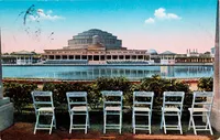 A group of six light (almost white) chairs standing in a neat row. In the background we can see a building with a dome, set against a blue sky, two small clouds visible. The building is the Centennial Hall and the chairs represent the Terrace Restaurant. This image is an old postcard. It’s a bit dirty and worn-out in the left corner.