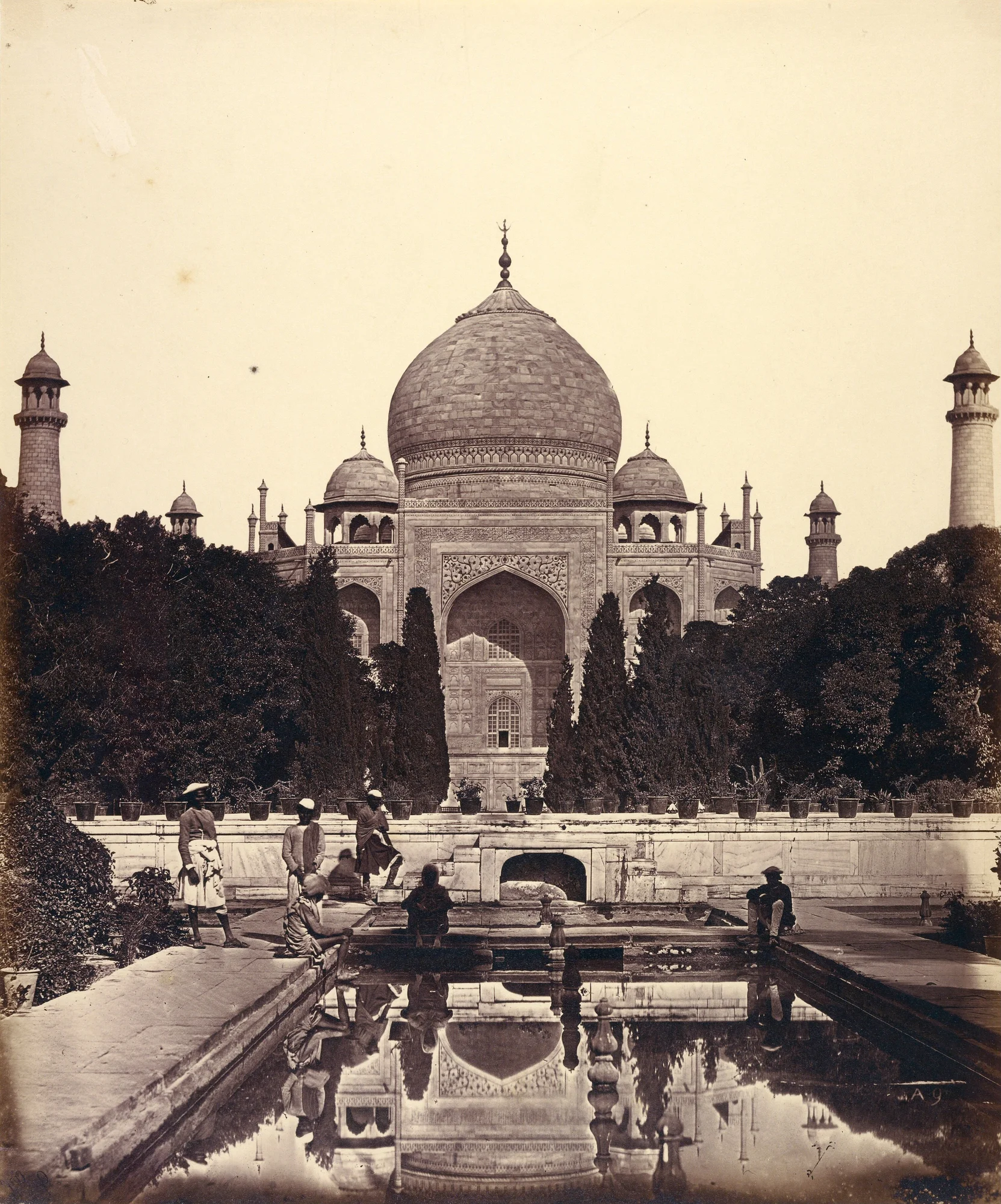 Black and white photograph of a large outdoor stone structure with rounded roofs, surrounded by trees, towers, and a large pool of water in the forefront.