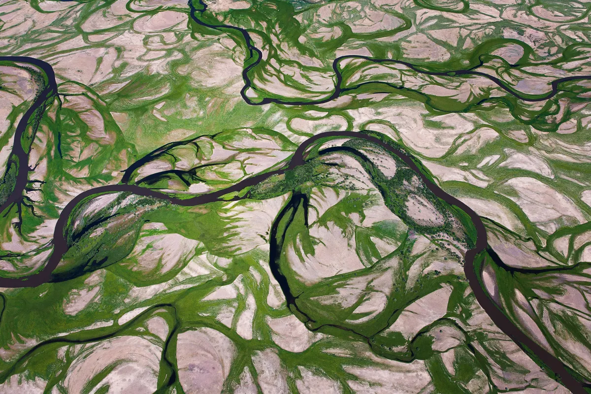 This image captures the intricate meanders of the Tula River in Töv, Mongolia, showcasing its winding path through a diverse landscape. The river is bordered by vibrant green vegetation, creating a striking contrast with the surrounding sandy and rocky ar