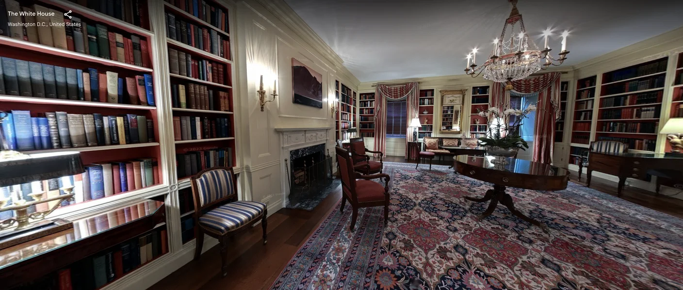A street view capture of the library located in the White House. On the floor is a rug with a burgundy and navy blue pattern. In the middle is a brown table and across are 2 red chairs with brown legs. In the middle of the table, there is a white floral arrangement. Shelves of books adorn the wall, and seating is found all around the room.