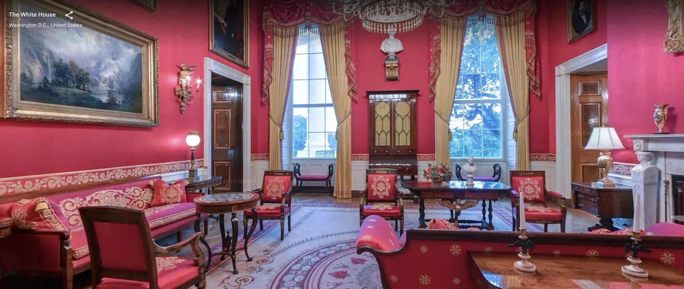 A street view capture of the Red Room in the White House. The walls are red as are the chairs. The are 5 red charis with brown wooden legs, accompanied by a red couch, 3 brown tables. There are 2 windows , each one adorned by red and gold curtains. There is a chandelier that’s on. The walls are adorned by portraits in gold frames.