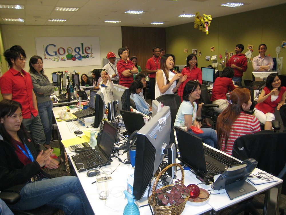 Three rows of people sitting and standing at desks with laptops and monitors inside an office with a Google sign on the wall.