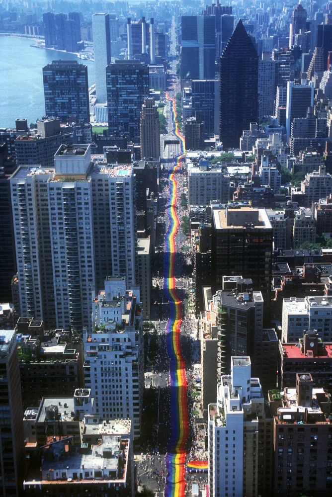 Bird perspective on a New York City street through which people are marching with a sevel-block long pride flag