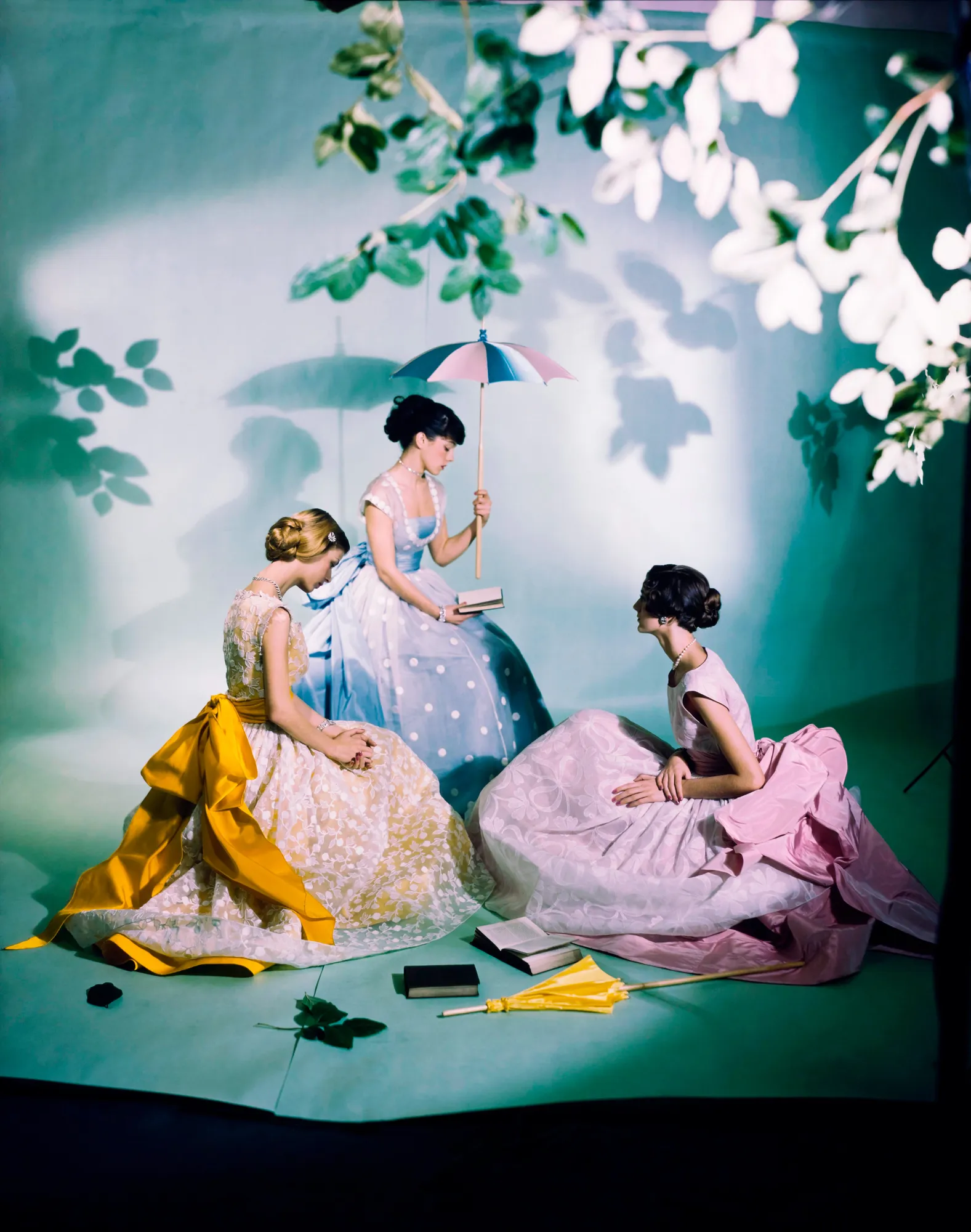 Three models wearing long colorful gowns, all looking away from the camera, sit in front of a white backdrop with flowering trees in the foreground