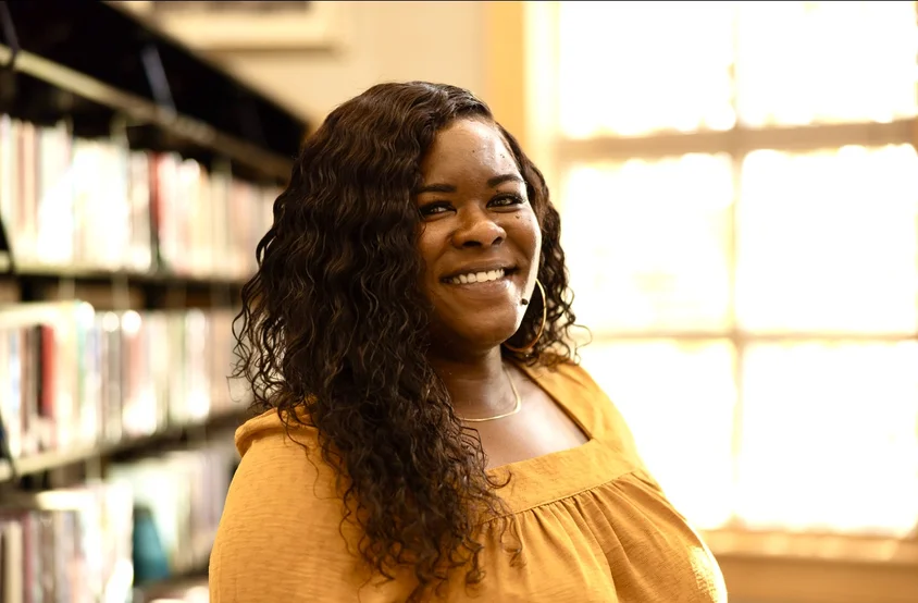 A woman wearing a yellow blouse stands next to a book shelf and smiles directly at the camerica.