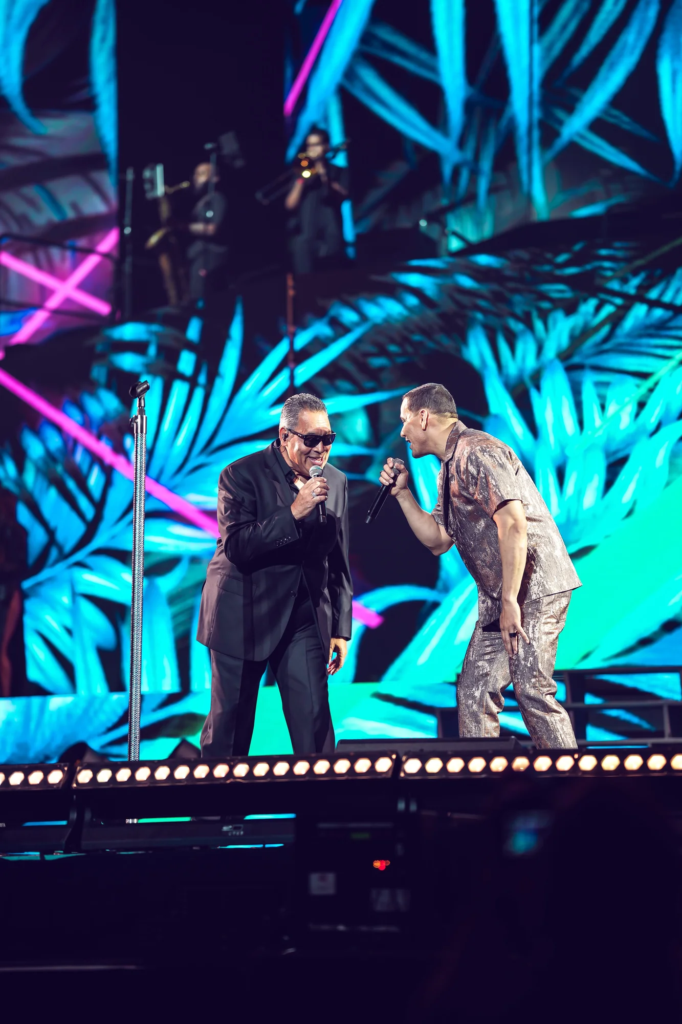 Victor Manuelle and Tito Nieves sing together into microphones on a stage. Victor Manuelle wears a silver two-piece outfit, while Tito Nieves wears a monochrome black suit with black sunglasses. Behind them is a screen with turquoise palm leaves and fuschia lines.