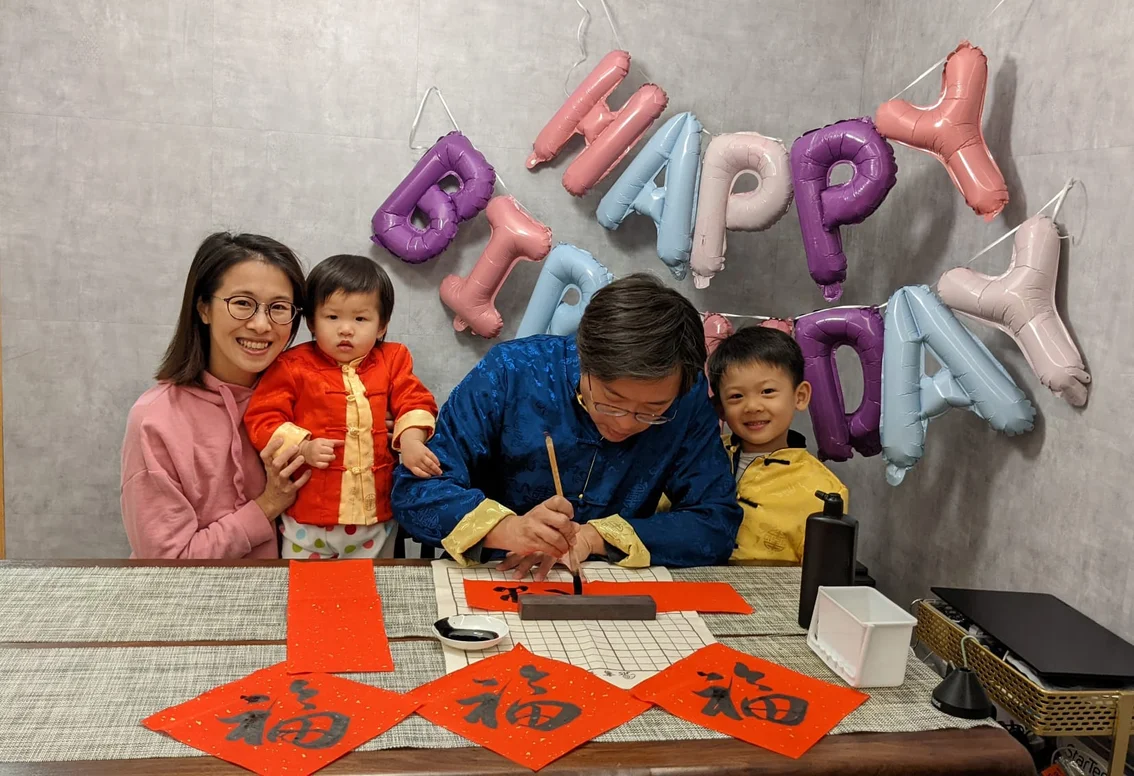 A photograph of a woman holding her small child and smiling into the camera. There is a happy birthday balloon sign in the background. Another small child looks into the camera and smiles while a father practices calligraphy.