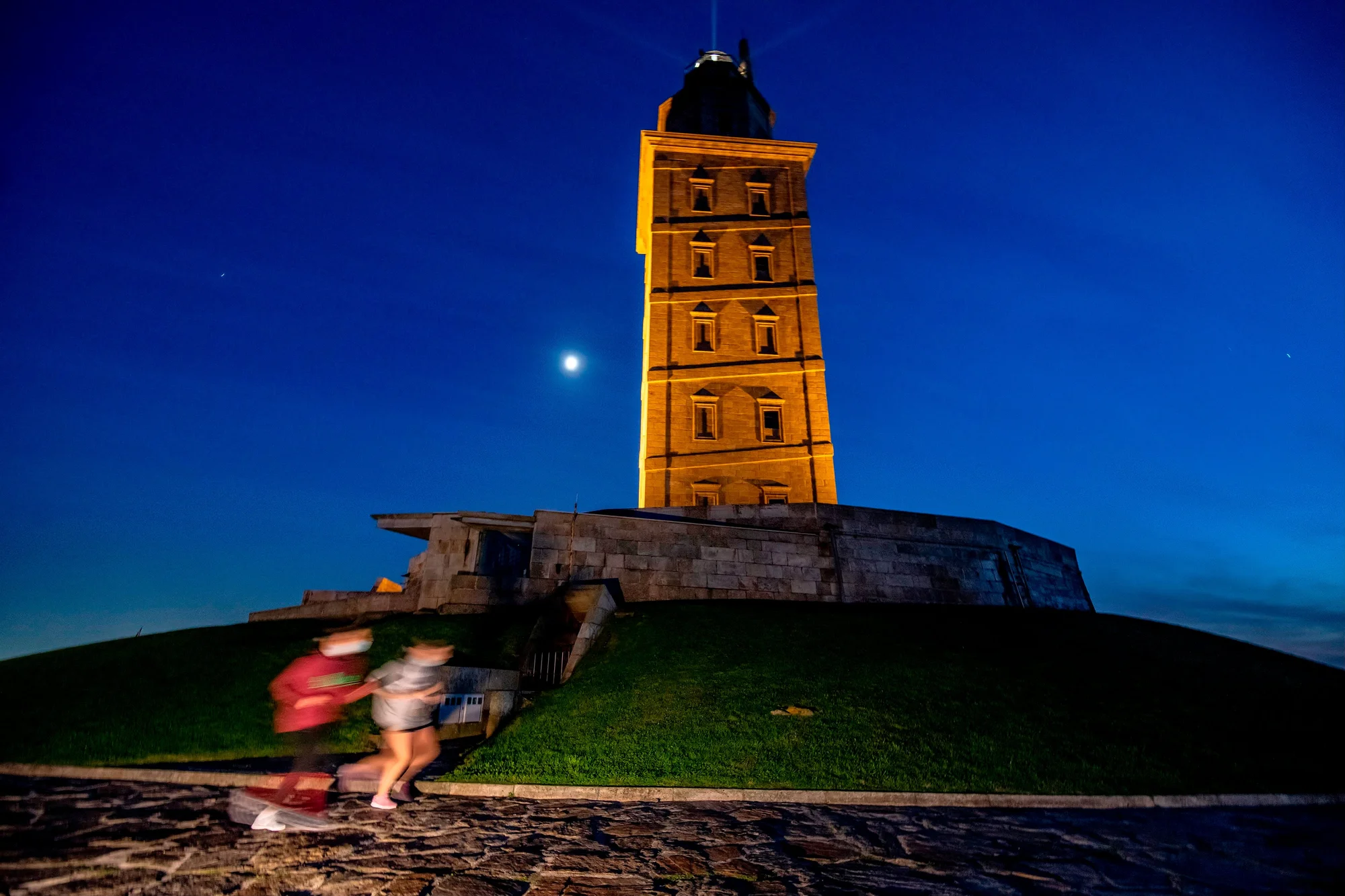 A photograph of the tower of hercules, an ancient Roman lighthouse, captured at night. Two blurred people appear in the foreground walking.