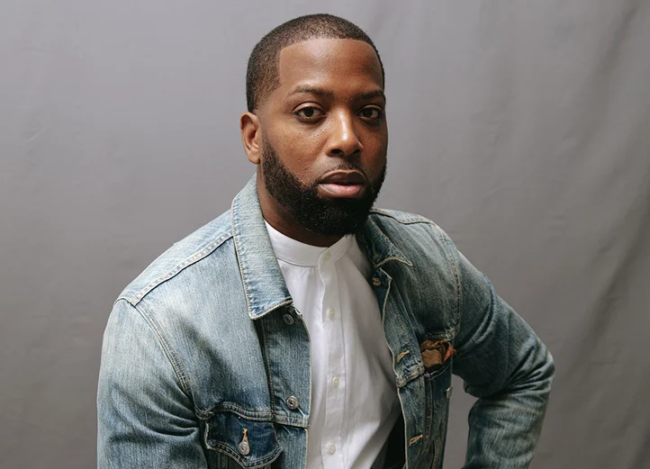 Tristan Walker looks seriously into the camera. He is wearing a white shirt and faded blue jeans jacket and is standing in front of a gray background.