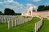 Rows of identical and equally spaced white, plain gravestones organized in circular arrangement with a memorial building, clouded blue sky and trees in the background.