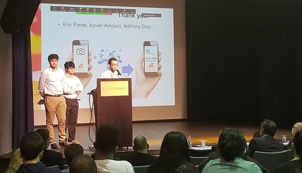 Eric, Xavier, and Anthony standing on stage behind Kean University podium and presenting their project to an audience.