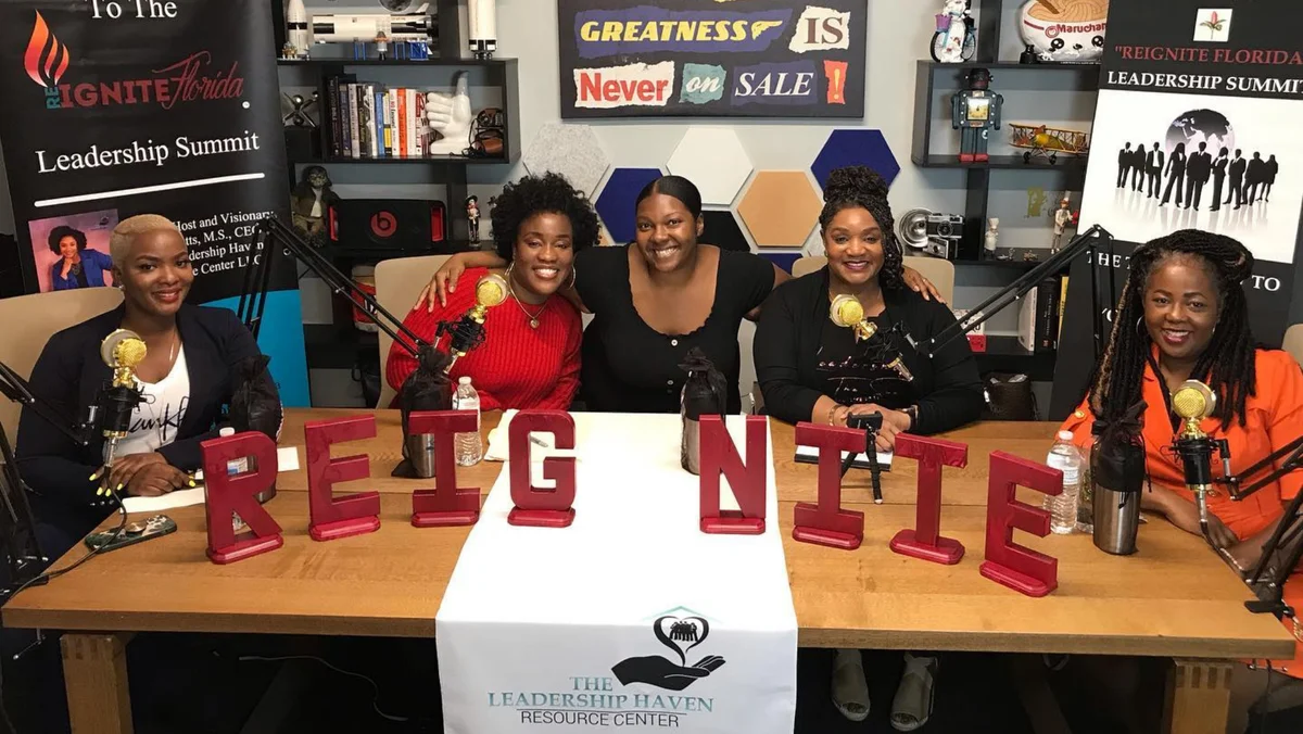 Five women sit at a table facing camera and smiling, with signs reading "Leadership Haven Resource Center" on both sides. Red letters reading the word "REIGNITE" are set up on table facing camera.