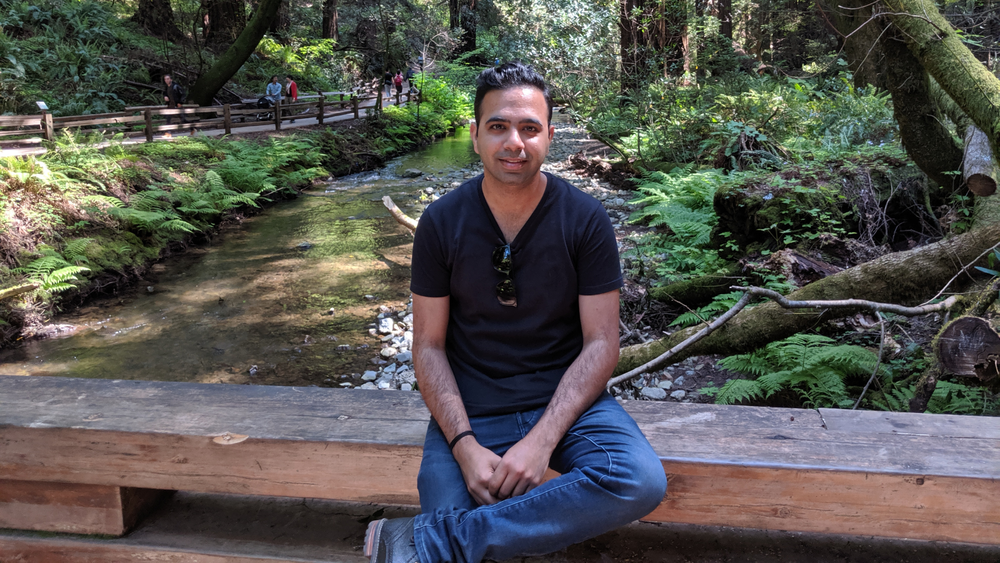 Sameer, in a black t-shirt and blue jeans, sits on a bridge overlooking a wooded river.