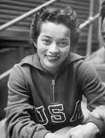 A black-and-white photograph of a woman sitting on a step with her hands placed on her knees. She is smiling and wearing a zip-up sweatshirt that reads “USA.”