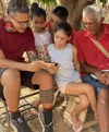 Three kids and an older man sit outside watching a man play a game on his phone