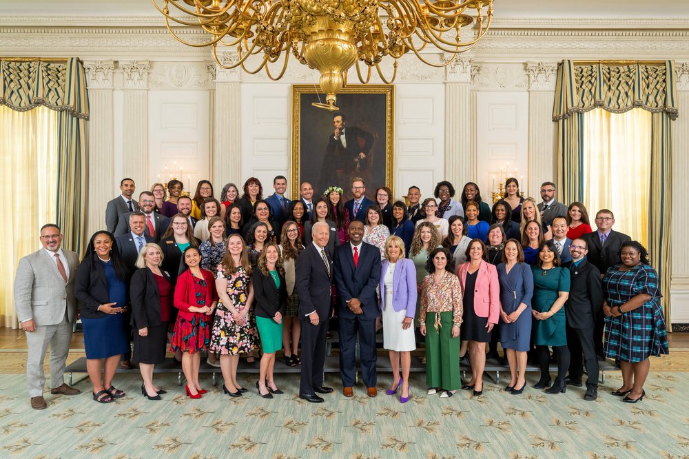 A large group of people — the 2022 State Teachers of the Year, President Joe Biden and First Lady Dr. Jill Biden — pose in front of a portrait of Abraham Lincoln. Above them is a golden chandelier.