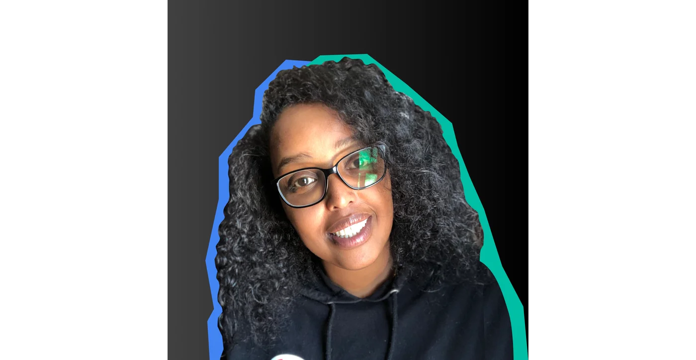 Image shows a woman with curly dark hair and glasses smiling at the camera against a black background.