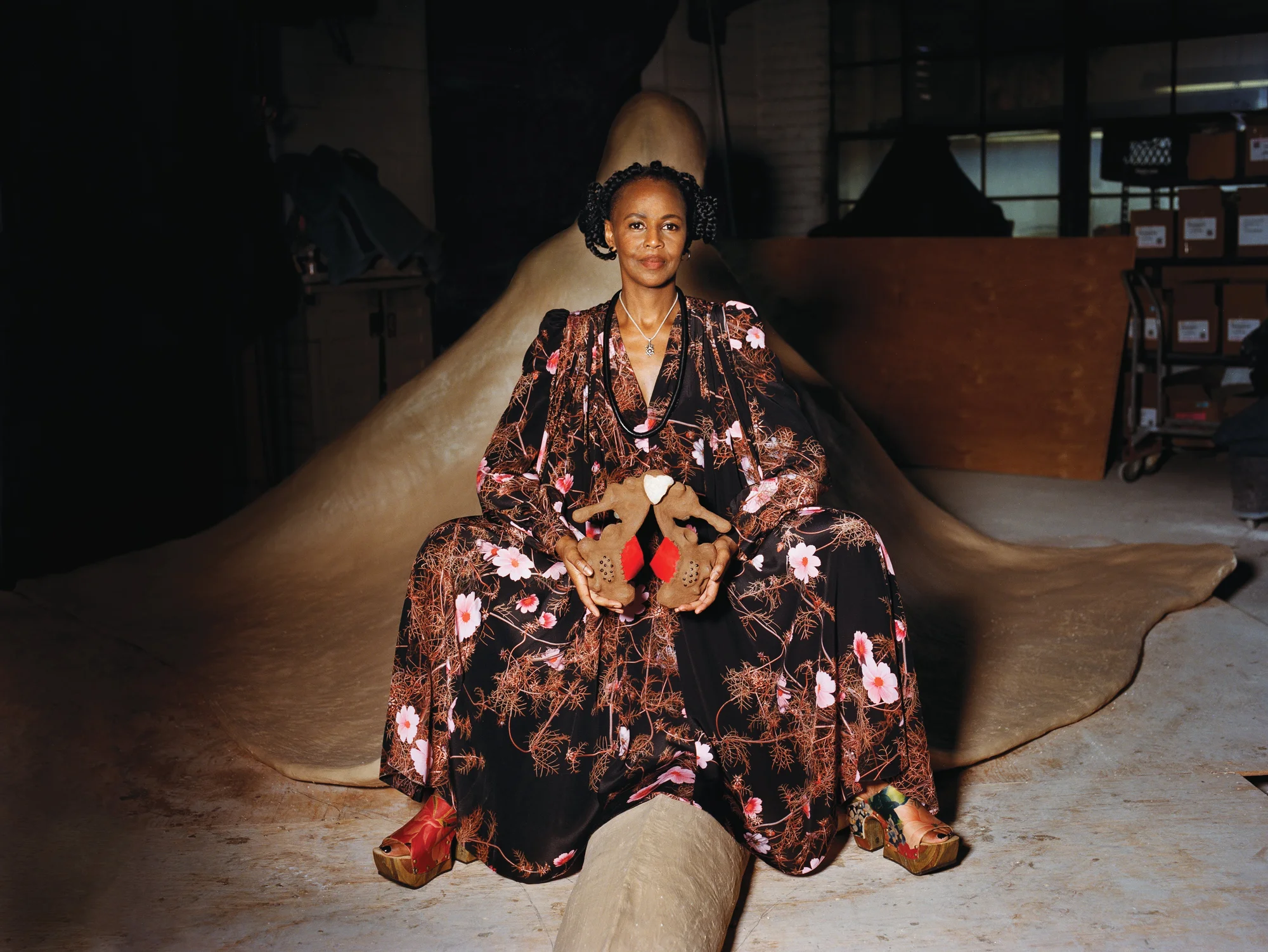 A woman sits on a large sculpture of a room-sized manta ray, and wears a long, black and pink patterned dress. She holds two smaller, seahorse-shaped sculptures; one in each hand. In the background is her workspace with various cardboard boxes and pieces of plywood.