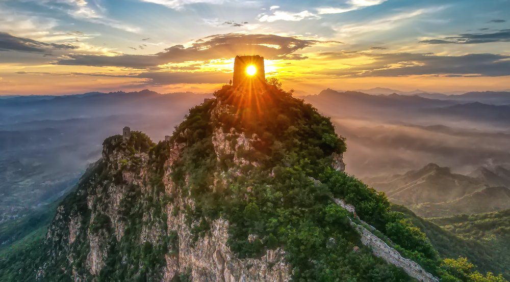 The 'Watching the Capital Tower', a tower on top of a wooded peak high above a sweeping mountain landscape, with the sun setting in the background.