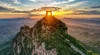 The 'Watching the Capital Tower', a tower on top of a wooded peak high above a sweeping mountain landscape, with the sun setting in the background.