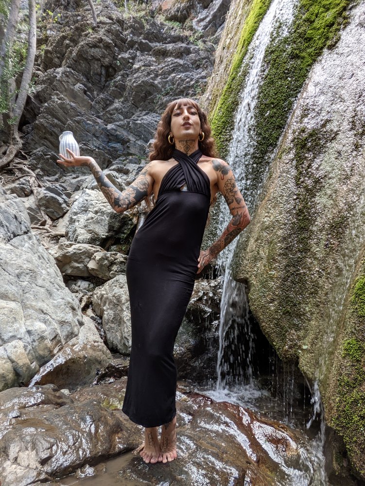 A person in a long black gown stands in front of a small water fall holding a crystal and looking into the camera.