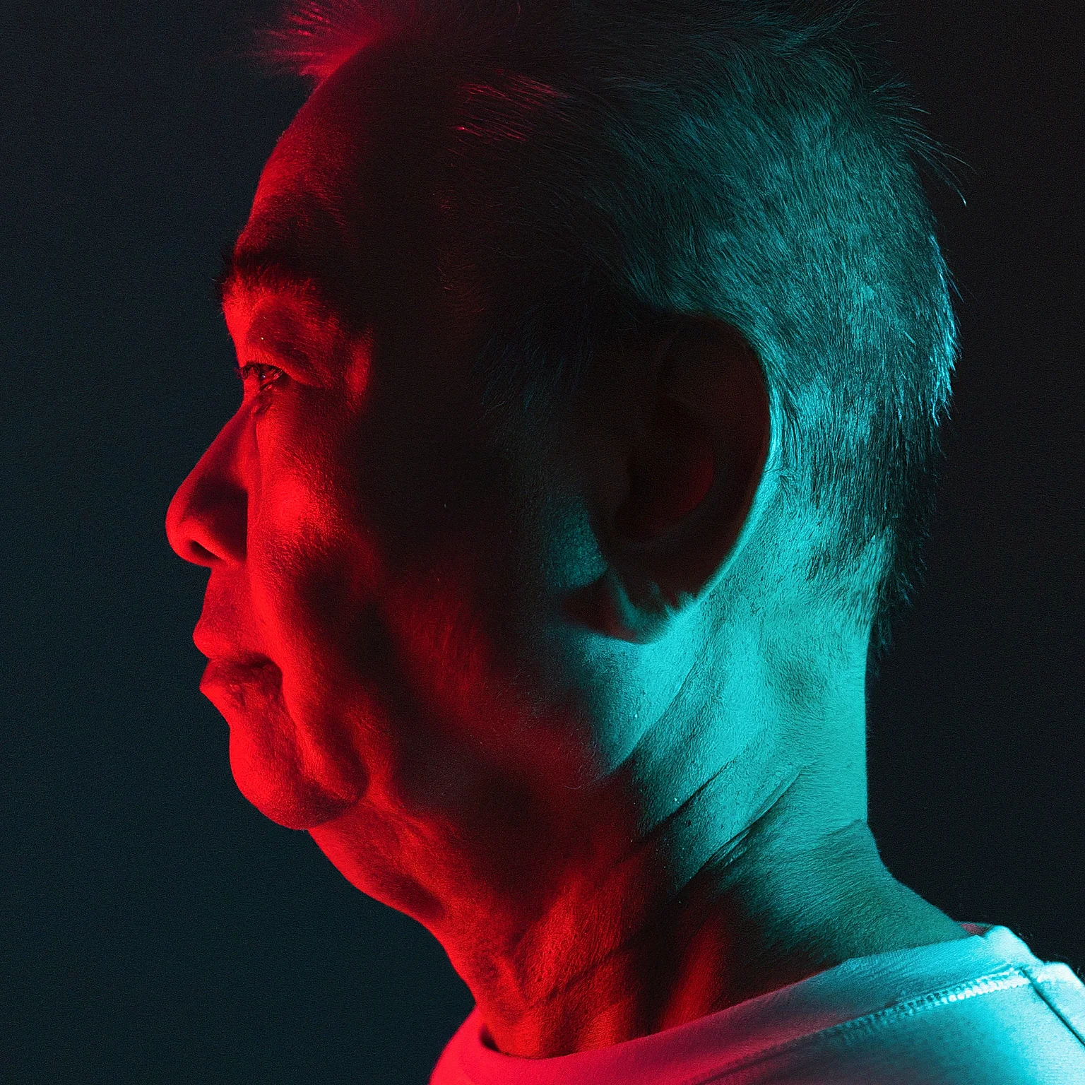 An image of an older man seen in profile, lit in front by red light and on the back of his head in teal light.