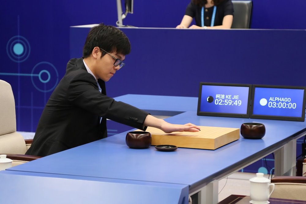 Grandmaster Ke Jie, wearing a suit and sitting on a white chair to the left of a blue table, leans forward to make a move on the board in his match against AlphaGo.