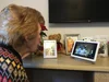 An elderly woman with brown hair wearing a sweater looks at Nest Hub Max screen that's playing a video of people tap dancing on it. The device is sitting on a table where she also has family photos in frames.