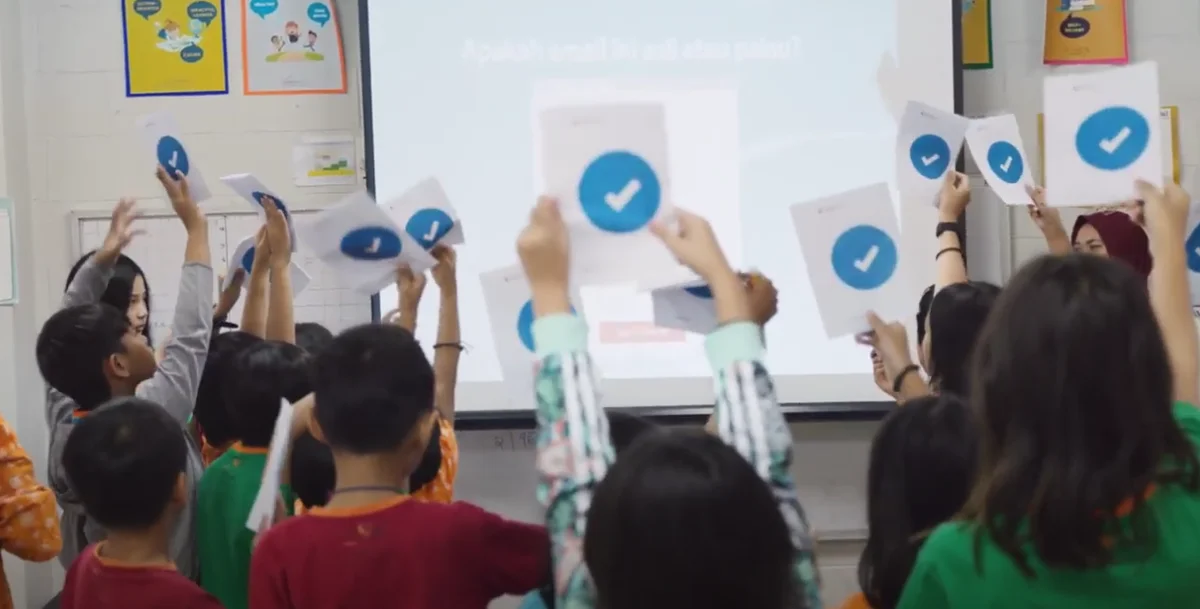 View from the back of a classroom of Indonesian schoolchildren. They are holding up signs with blue ticks on them, as part of the 'Be Internet Awesome' online safety course. A whiteboard and teacher are visible at the front of the class.