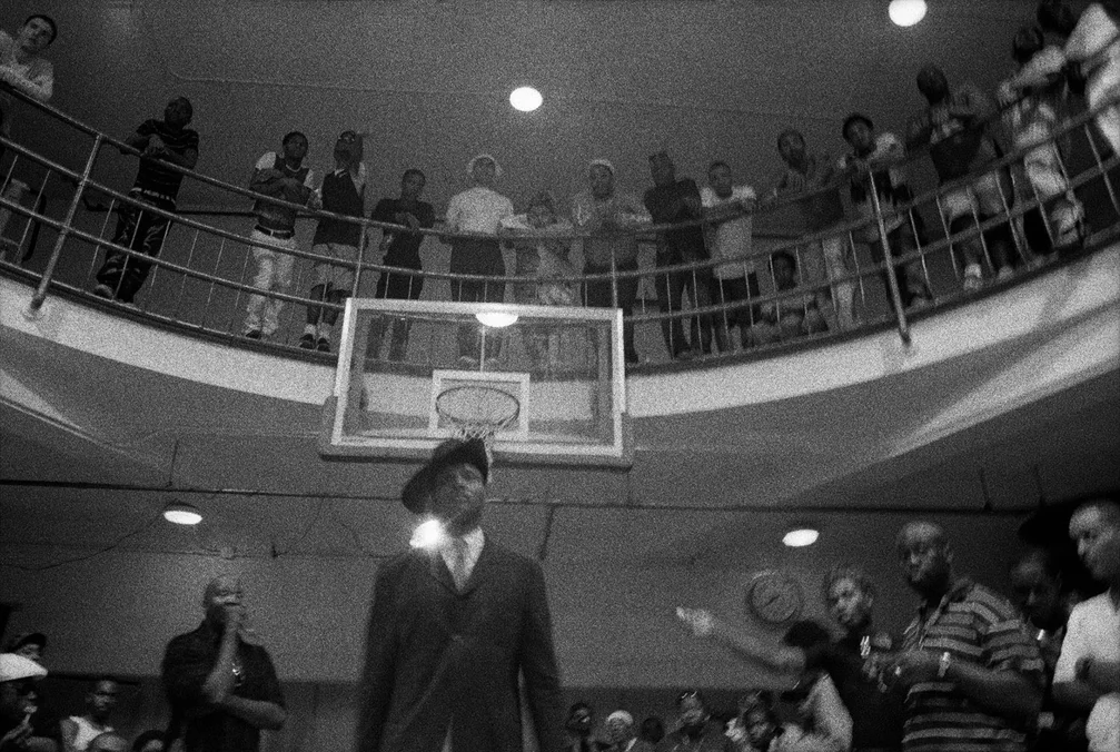 A large group of people are standing in a semicircle on two floors within a YMCA building, in front of a basketball hoop. The person most prominent in the foreground looks serious and powerful while wearing a suit and hat.