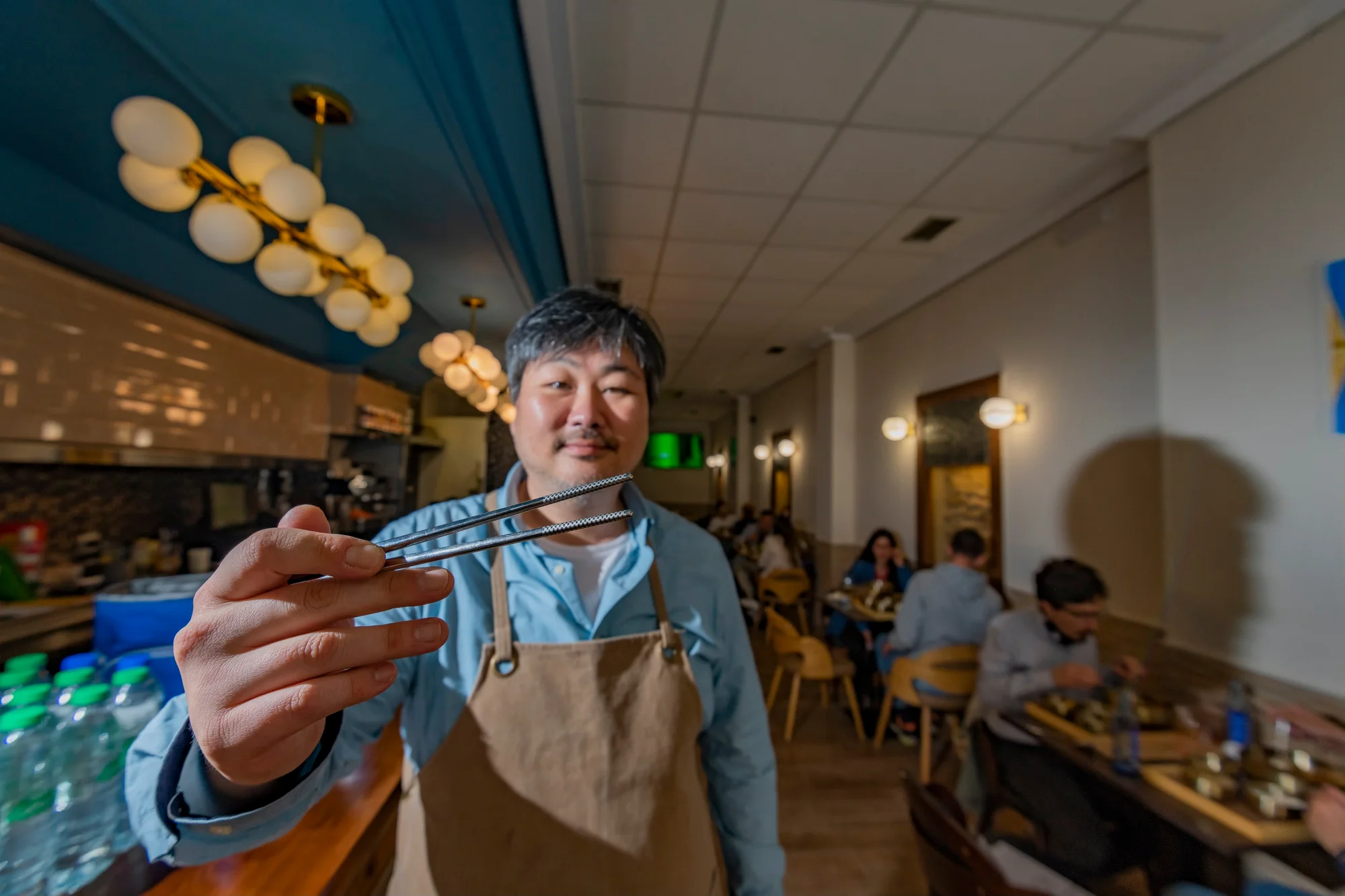 Photograph of a person of Asian origin holding chopsticks. The photograph was taken in a restaurant, in the background there are some tables.