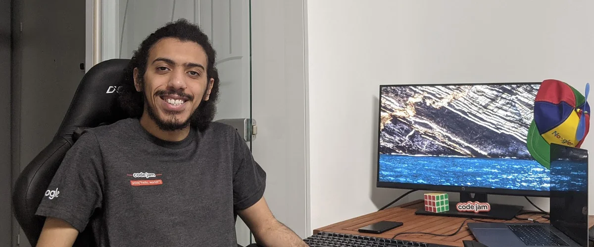 Article's hero Yosri sitting at his desk at home while wearing a Code Jam shirt. On the desk are a monitor, a laptop, a phone, a Rubix cube and a Noogler hat.media