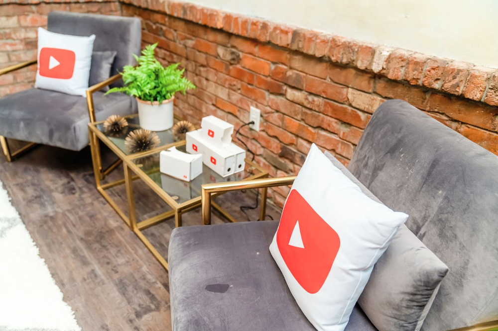 Two unoccupied grey chairs with white pillows featuring YouTube logos, either side of a pair of glass-topped tables with plants and YouTube-branded hardware boxes.