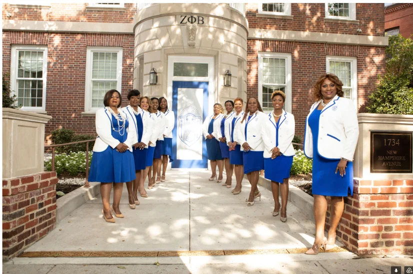 A group of women wearing blue dresses and white jackets standing outside a building.