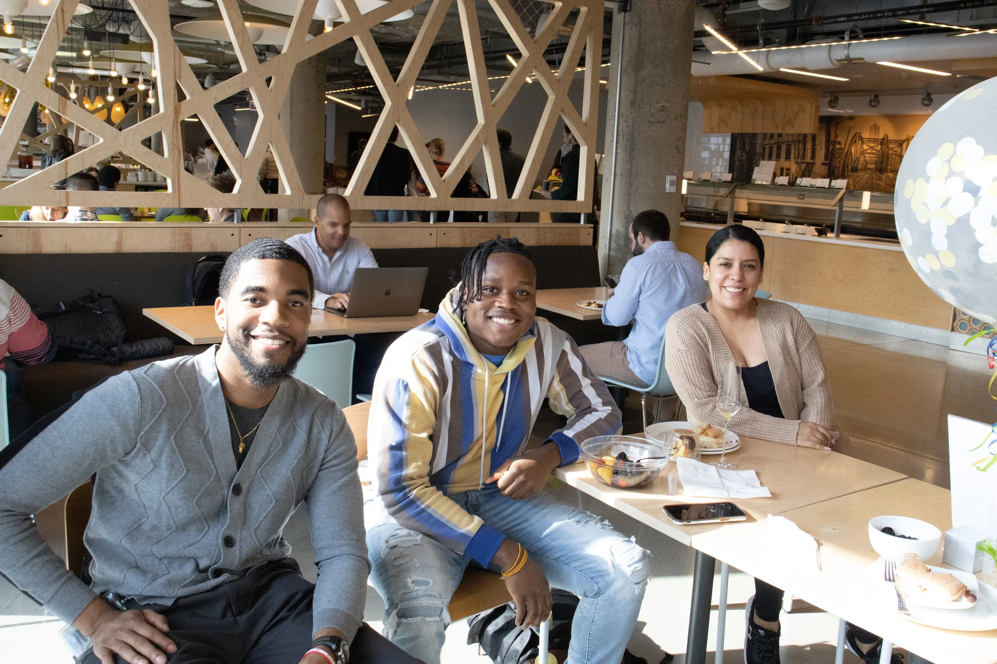 Three people sit at a table and smile at the camera. From left to right is Daniel, George and Yaseline. Daniel is wearing a gray cardigan with blue jeans, George is wearing a blue and yellow striped shirt and blue jeans, and Yaseline is wearing a light brown cardigan and a black tank top.