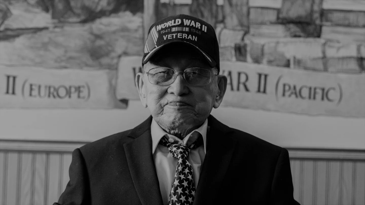 A black-and-white photograph of a man wearing glasses, a star-patterned tie and a hat that reads “World War II 1941 - 1945 Veteran.”