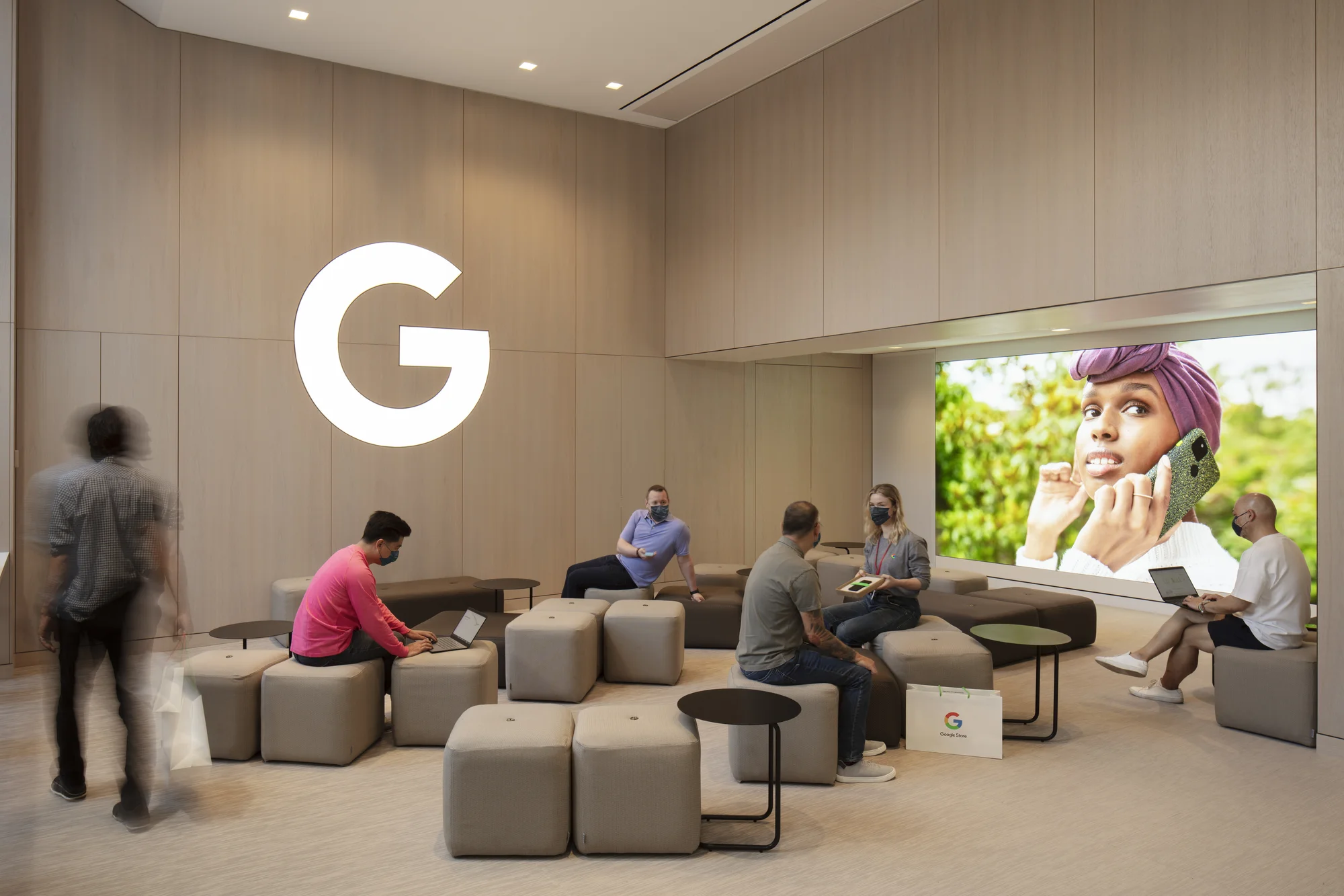 Six people stand and sit in a large meeting space containing a big screen and a Google ‘G’.