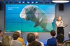 Woman standing in front of a screen with a dugong on it. Text on screen says “You and me, we need seagrass.”