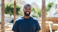 Prompt: A photo of a man with short hair and beard smiling at the camera. The background is blurry and it shows trees and buildings in light colors.