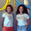 Two Black women with curly brown hair and wearing white t-shirts stand in front of a colorful mural.