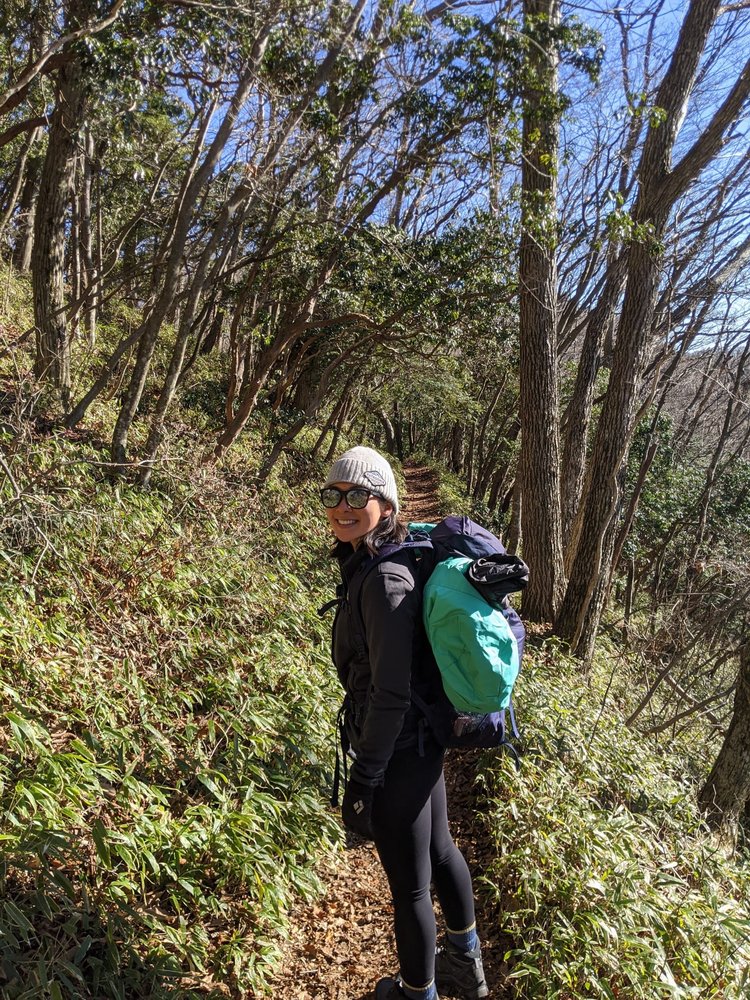 Eri Shinose in dark hiking gear, sunglasses and a white beanie, wearing a green backpack and smiling over her shoulder towards the camera. She's on a trail through a wooded hillside.